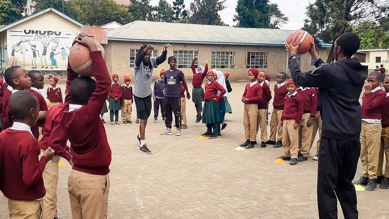 Schüler des Basketballprojekts in Tansania schauen zwei Trainern dabei zu, wie diese die Wurftechnik erklären.