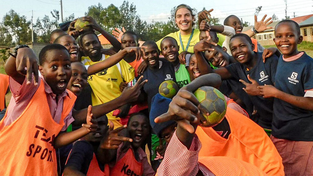 Ein Sporttrainer beim Freiwilligendienst im Ausland zusammen mit einer Gruppe feiernder Kinder und Jugendlicher in Sporttrikots.