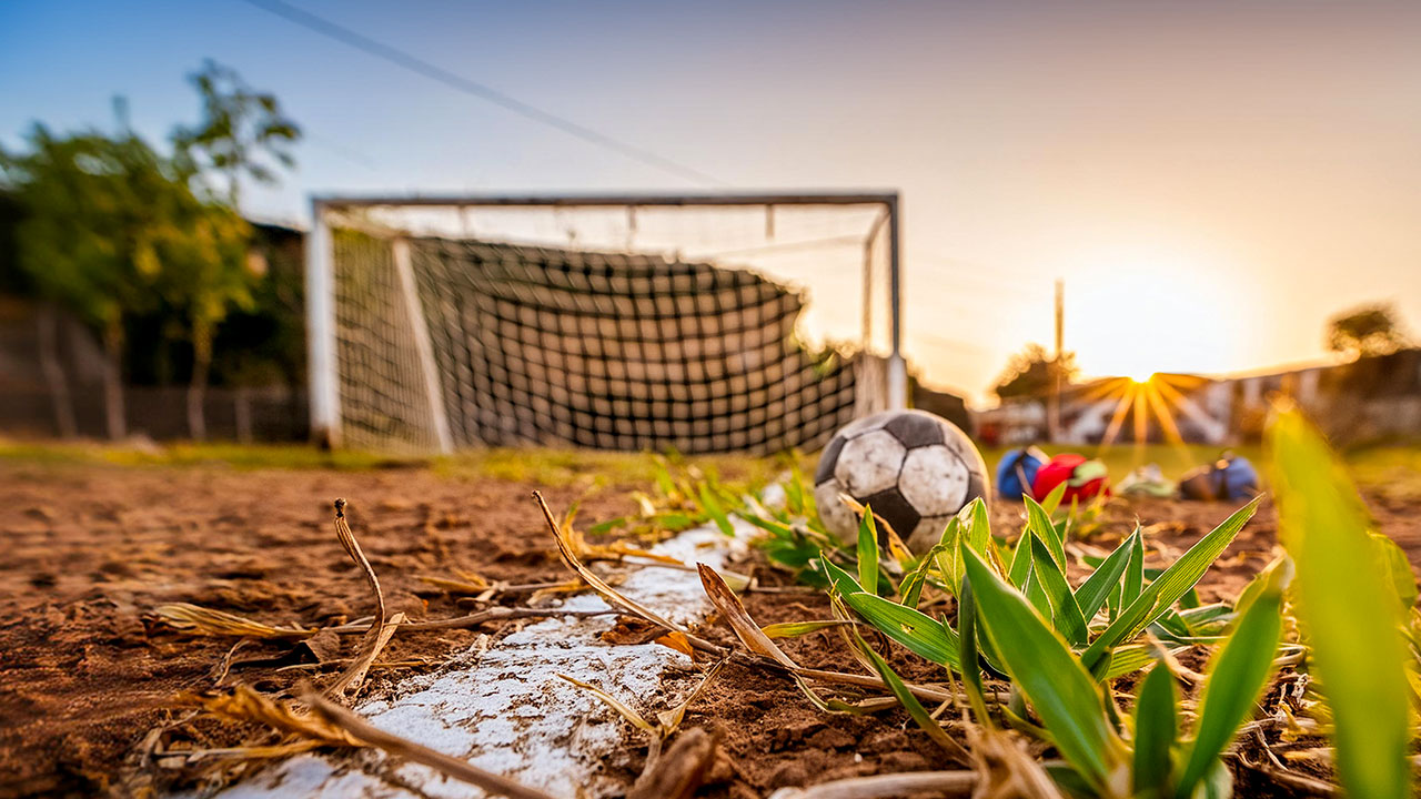 Nahaufnahme eines trockenen Fußballplatzes mit einem Fußball vor einem verwittertem Tor bei Sonnenuntergang.