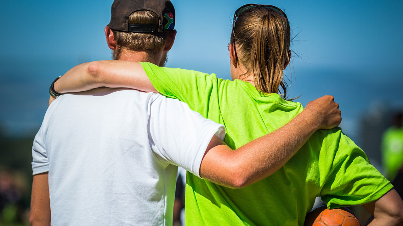 Two volunteer sports coaches have their arms around each other's shoulders, watching the exercise.