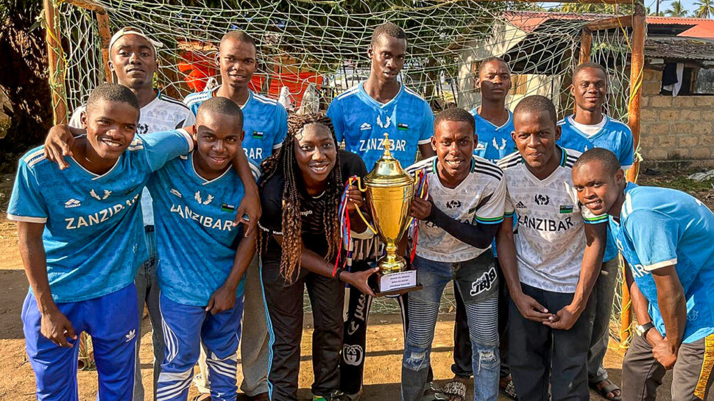 The coach poses with a boys’ team in front of a handball goal, holding a trophy.