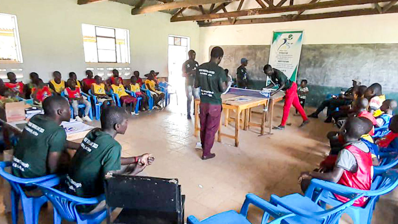 Young students and athletes from a Kenyan school sit on chairs in a circle in a classroom, listening to the coach's explanations.