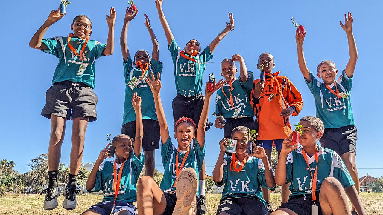 Children from a sports team leap into the air with their hands in the air, holding their trophies.