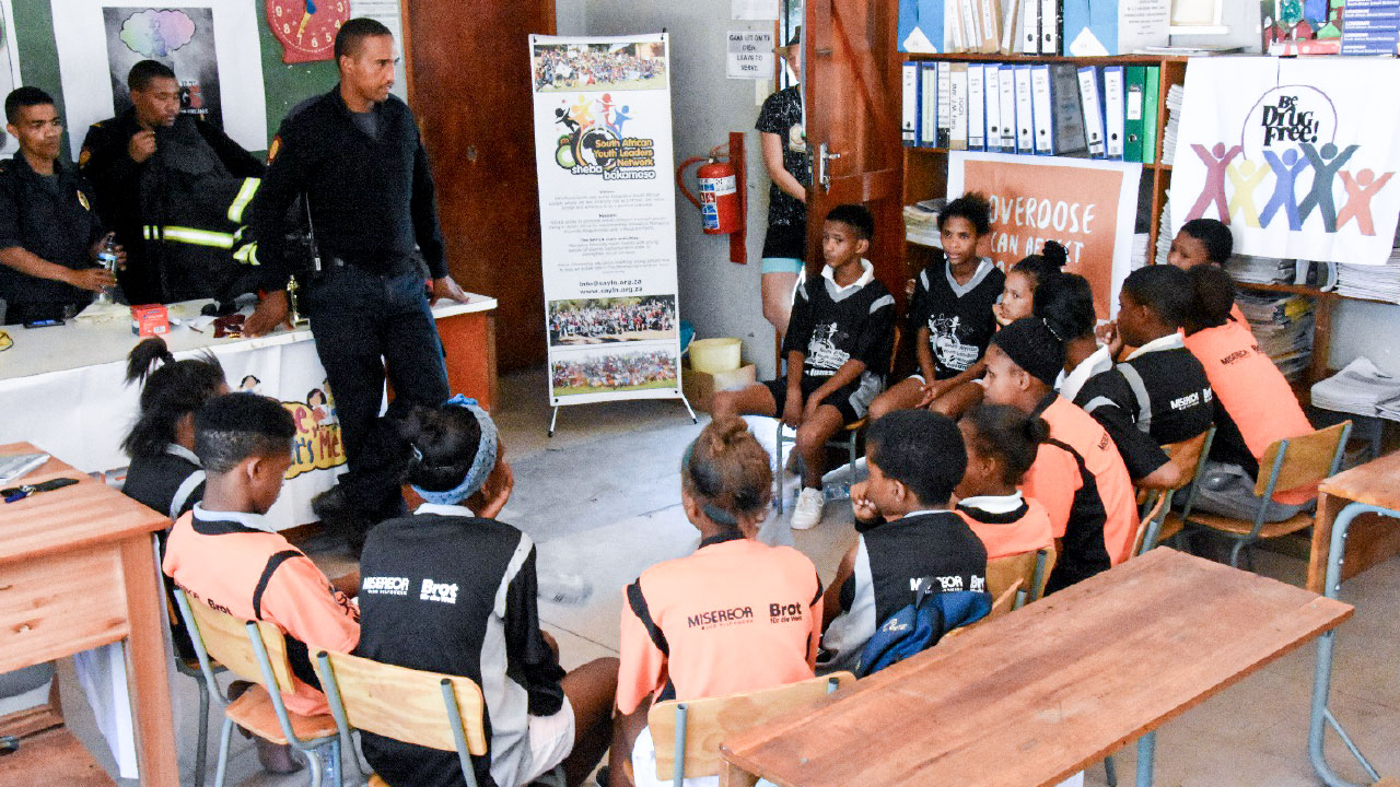 Junge Schüler einer Schulklasse in Südafrika sitzen mit Männern in Polizeiuniformen im Kreis im Klassenzimmer und werden über Drogenmissbrauch aufgeklärt.