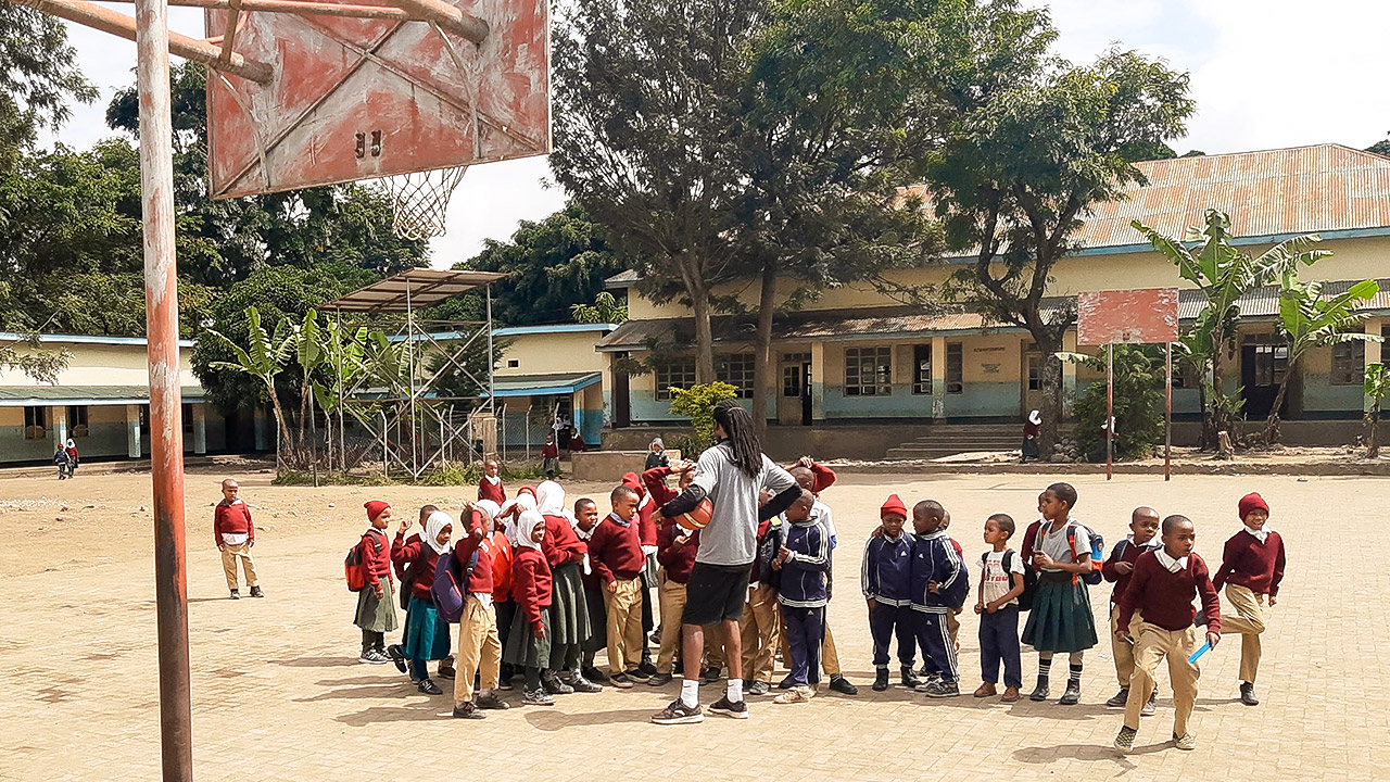 Auf einem sandigen Schulhof in Tansania hören Jungen und Mädchen aufmerksam dem Basketballtrainer zu.