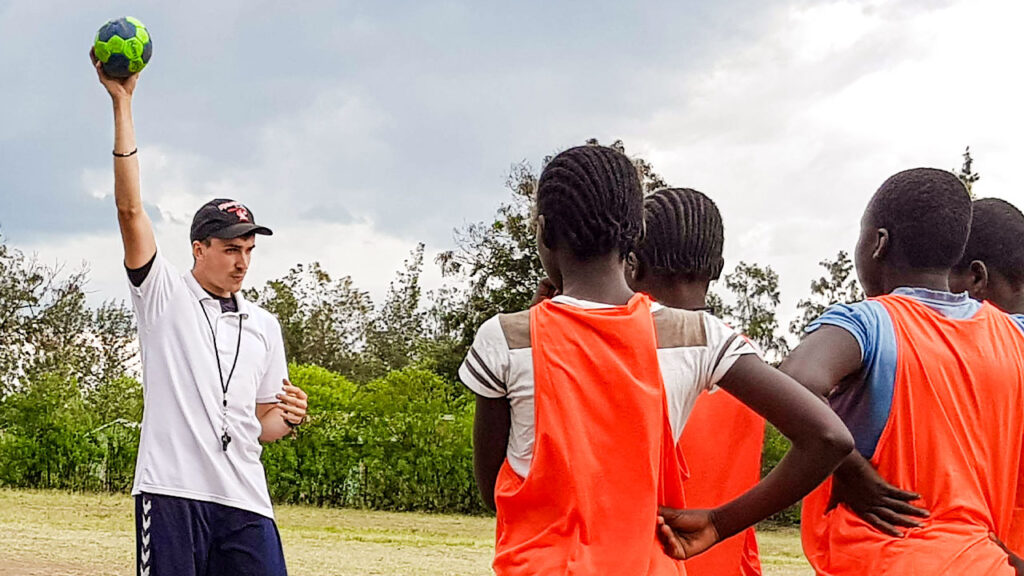 Ein Sporttrainer unterrichtet beim Freiwilligendienst im Ausland eine Gruppe Mädchen und hält dabei einen Handball hoch.