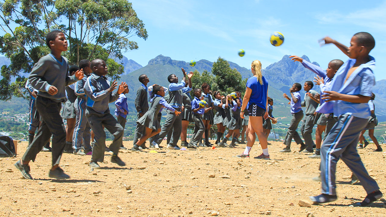 Under the watchful eye of a sports volunteer, children in school uniforms practise throwing the ball on a sandy and stony surface.