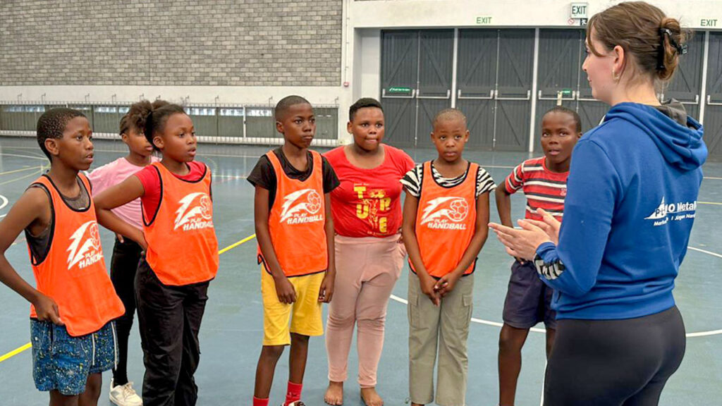 Während ihres Freiwilligendienstes im Ausland erklärt eine Sportvolontärin einer Gruppe von Jungen und Mädchen in der Sporthalle einer Schule die Taktik des Handballspiels.