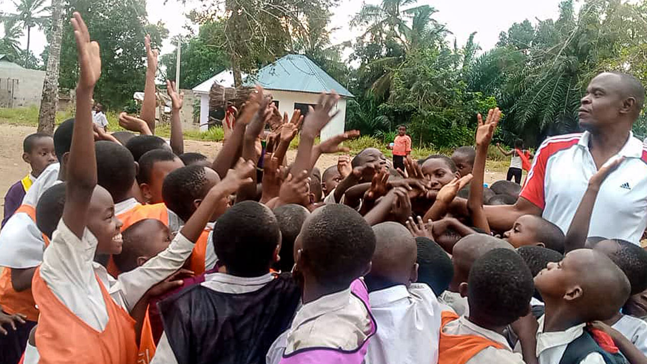 A large group of schoolchildren stand tightly packed around the sports coach, smiling and raising their hands in joy.