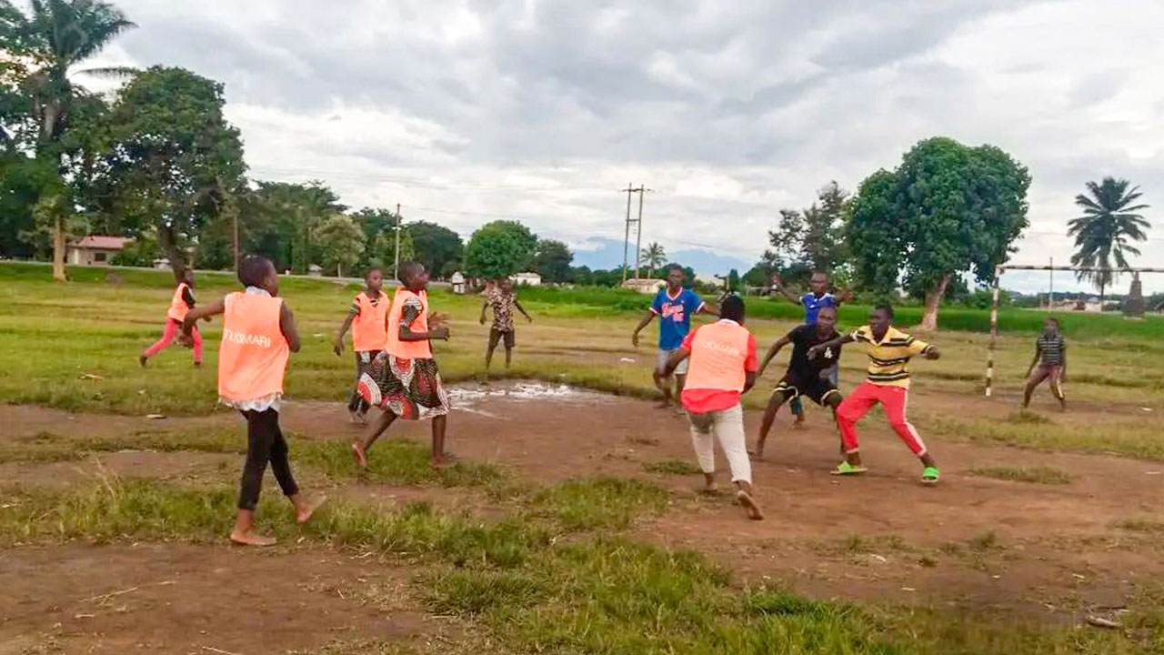 Barefoot and wearing loose sportswear, young people from a village in Tanzania play handball on an uneven grass surface with a goal without a net.